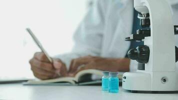 Medical worker in lab coat and sterile mask, doing a microscope analysis while her colleague are working behind video