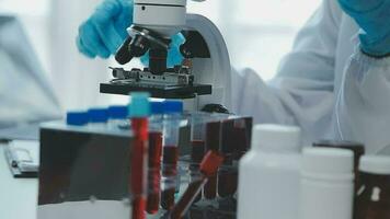 Medical worker in lab coat and sterile mask, doing a microscope analysis while her colleague are working behind video