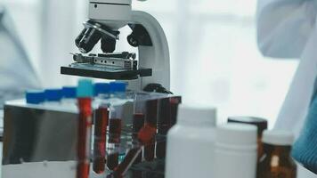 Medical worker in lab coat and sterile mask, doing a microscope analysis while her colleague are working behind video