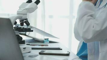 Medical worker in lab coat and sterile mask, doing a microscope analysis while her colleague are working behind video
