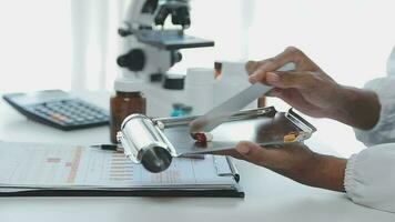 Medical worker in lab coat and sterile mask, doing a microscope analysis while her colleague are working behind video