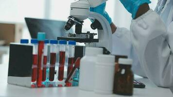 Medical worker in lab coat and sterile mask, doing a microscope analysis while her colleague are working behind video