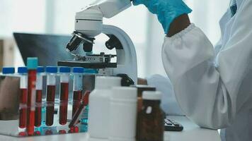 Medical worker in lab coat and sterile mask, doing a microscope analysis while her colleague are working behind video