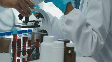 Medical worker in lab coat and sterile mask, doing a microscope analysis while her colleague are working behind video