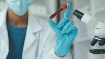 Medical worker in lab coat and sterile mask, doing a microscope analysis while her colleague are working behind video