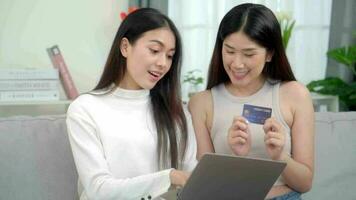 twee Aziatisch Dames gebruik makend van online boodschappen doen credit kaarten via hun laptops Aan sofa Bij huis. online succes kopen uitverkoop. video