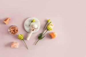 A professional rose quartz massager for facial care at home lies on a white marble round podium and sea salt in a bowl. Top view. austoma flowers photo