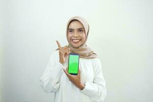 Excited beautiful Asian muslim woman showing green screen mobile phone isolated over white background photo