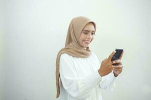 hermosa sonriente asiático musulmán mujer vistiendo hijab participación móvil teléfono en mano aislado en blanco antecedentes estudio retrato. personas religioso estilo de vida concepto foto