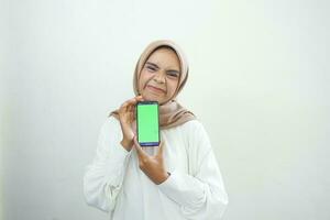 Excited beautiful Asian muslim woman showing green screen mobile phone isolated over white background photo