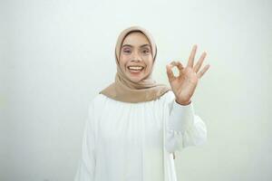 Smiling young Asian woman in white casual showing okay gesture demonstrates symbol of approval isolated over white background photo