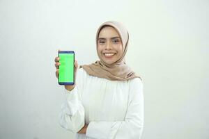 Excited beautiful Asian muslim woman showing green screen mobile phone isolated over white background photo