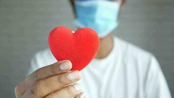 a man in a white shirt and surgical mask holds a red heart video