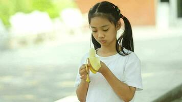 pequeno ásia menina comendo banana dentro a parque. seletivo foco. video