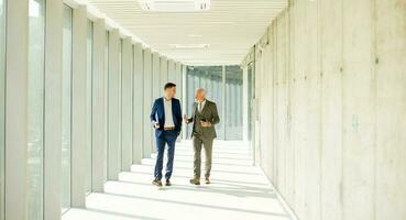 Young and a senior businessman walk down an office hallway, deep in conversation photo