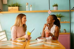 Young black and caucasian woman having good time, drinking fresh juices and having healthy breakfast in the cafe photo