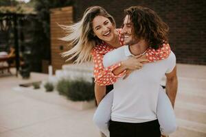 Handsome long hair man carrying the young woman on his back in front of brick house photo