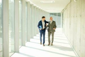 Young and a senior businessman walk down an office hallway, deep in conversation photo