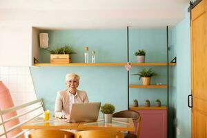 Senior woman using mobile phone while working on laptop and drinking fresh orange juice in the cafe photo