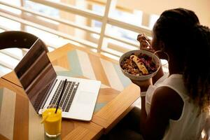 Young black woman having a healthy breakfast while working on laptop in the cafe photo