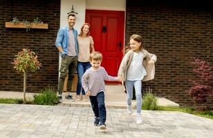 Mother and father standing outside on the front porch of a brick house, while son and daughter running photo