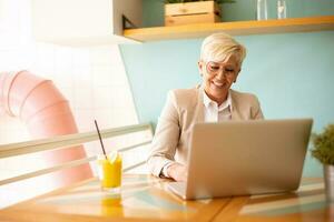 Senior woman using mobile phone while working on laptop and drinking fresh orange juice in the cafe photo