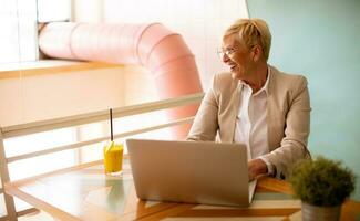 Senior woman using mobile phone while working on laptop and drinking fresh orange juice in the cafe photo