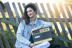 A beautiful country girl in bright clothes sits on a wooden bench with a cassette recorder. Woman in the style of the 90s. photo