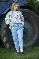 A beautiful country girl in the style of the 90s in bright clothes stands near a tractor. photo