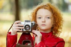 Little red-haired girl with a retro camera on an autumn background.Little red-haired girl with a retro camera on an autumn background.Close-up of the girl's face with a vintage camera. photo