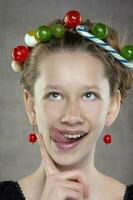 A funny teenager girl with a wreath of sweets on her head shows her tongue. photo