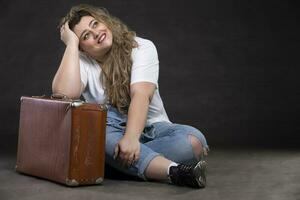 Beautiful fat woman in denim clothes with an old retro suitcase on a gray background. Plus size model girl. photo