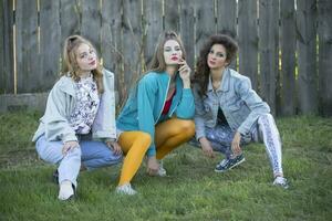 Funny girls in the style of the 90s in bright clothes are sitting near a wooden fence. photo