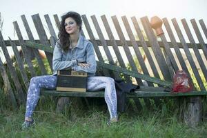 un hermosa país niña en brillante ropa se sienta en un de madera banco con un casete grabadora. mujer en el estilo de el años 90 foto