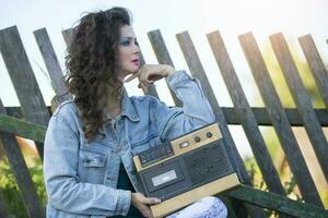 A beautiful country girl in bright clothes sits on a wooden bench with a cassette recorder. Woman in the style of the 90s. photo