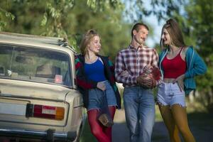 Handsome girls and a country man dressed in 90s style stand near an old car. photo