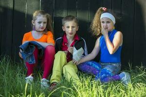 Beautiful model children in bright clothes sit on the grass near a wooden fence. photo