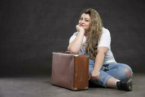 Beautiful fat woman in denim clothes with an old retro suitcase on a gray background. Plus size model girl. photo