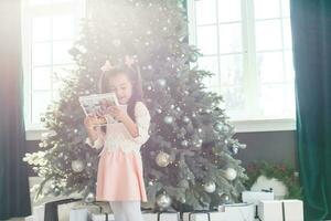 pequeño niña mirando fotolibro en frente de Navidad árbol foto