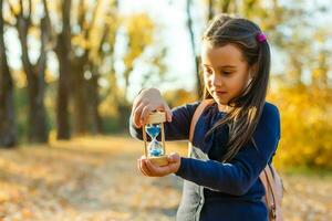 pequeño niña Chica de escuela sostiene reloj de arena en otoño foto