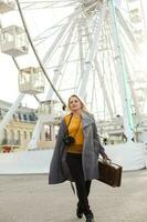 Attractive blonde girl in casual clothing, wearing a coat, holding a brown retro suitcase in his hands, background of the ferris wheel photo