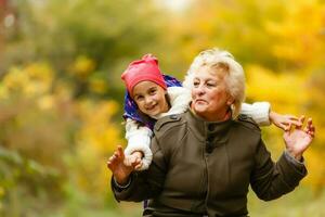 contento mayor dama y un pequeño niñito chica, abuela y nieta, disfrutando un caminar en el parque. niño y abuelo. otoños día. abuela y pequeño niña contento juntos en el jardín foto