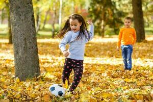 pequeño niña pateando pelota en el otoño parque foto
