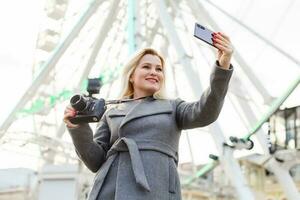 Attractive blonde girl in casual clothing, wearing a coat, takes selfie, looking into the camera. Stylish girl takes selfie, background of the ferris wheel photo