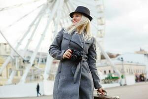Attractive blonde girl in casual clothing, wearing a coat, holding a brown retro suitcase in his hands, background of the ferris wheel photo