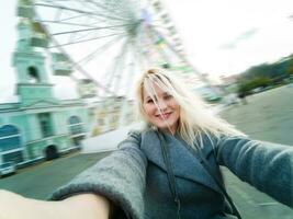 Attractive blonde girl in casual clothing, wearing a coat, takes selfie, looking into the camera. Stylish girl takes selfie, background of the ferris wheel photo