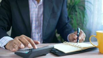 une homme dans une costume est l'écriture sur une carnet et en utilisant une calculatrice video