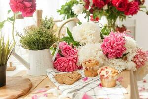 The kitchen countertop is decorated with peonies. The interior is decorated with spring flowers. Pink peonies and sweet cupcakes on a wooden countertop. Interior details. photo