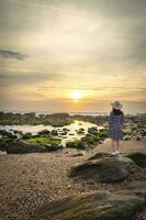 mujer con sombrero disfrutar el puesta de sol a el rocoso playa en gaia, Oporto, Portugal. foto