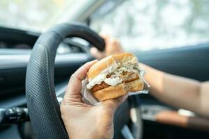 asiático dama participación hamburguesa a comer en auto, peligroso y riesgo un accidente. foto